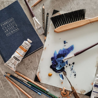 Art supplies including a Fabriano artist's journal, paint tubes, brushes, and a palette with blue paint on a textured surface. Photo from Laura J Stratton's art studio.