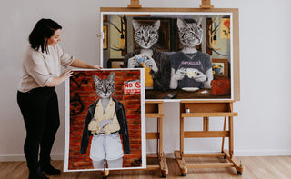 Artist Laura J Stratton standing with two of her artworks that show cat-ladies, feline heads on female bodies.