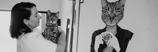 Artist Laura J Stratton holding her cat in her studio. Standing next to her realistic artwork showing a cat with a woman's body holding a teacup.