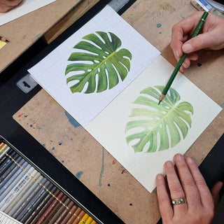 Person drawing a green leaf with a pencil on a piece of paper, surrounded by art supplies.