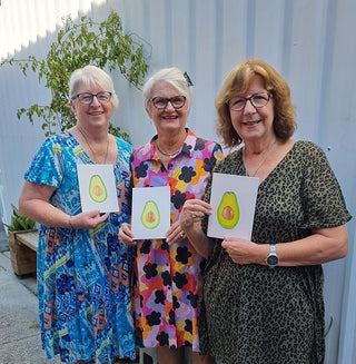 Three women holding drawings of avocados in front of a white wall.